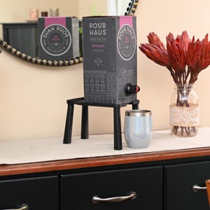 May include: A black wine-on-tap box labeled "Pour Haus Red Blend American" sits on a small black stand. A light blue tumbler and a vase with red flowers are next to it. A mirror with a decorative border is in the background.