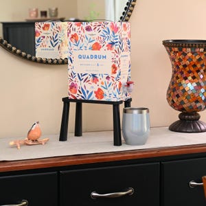 Puede incluir: Una caja de vino con estampado floral, etiquetada "QUADRUM", con un grifo rojo, se encuentra sobre un soporte negro. Un pequeño vaso aislado azul claro y un jarrón decorativo de mosaico están cerca. Una pequeña figura de pájaro en una rama.