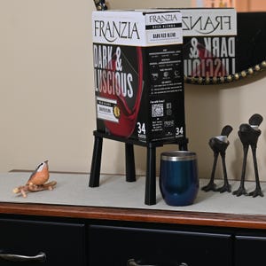 Könnte beinhalten: Eine schwarze Box mit Franzia Dark & Luscious Bold Red Blend Wein auf einem schwarzen Metallständer. Die Box steht auf einem Holztisch mit einer kleinen Vogelfigur und einem blauen Becher.