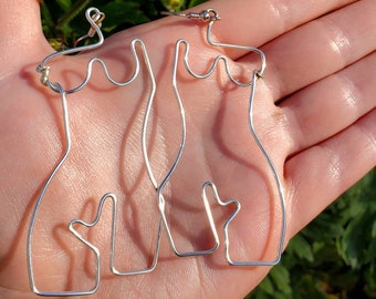 Pode incluir: Brincos de arame prateado em forma de figura feminina. Os brincos são segurados em uma mão aberta, com flores amarelas e folhagem verde ao fundo. Os brincos têm um design minimalista.