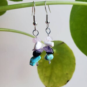 May include: A pair of dangle earrings with silver-colored hooks. Each earring features a stack of colorful gemstones, including pink, white, purple, black, and turquoise. The earrings are suspended against a blurred green background.