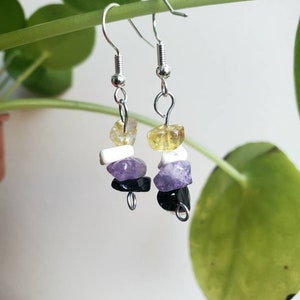 May include: Dangle earrings with silver hooks, each featuring a stack of gemstones: yellow, white, purple, and black. The earrings hang from a green stem, with a blurred background. Gemstones are measured in millimetres.