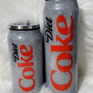 Puede incluir: Dos vasos de viaje con forma de lata de plata con purpurina con texto negro que dice "Diet Coke" y texto rojo que dice "Coke".