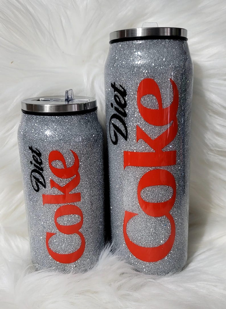 Puede incluir: Dos vasos de viaje con forma de lata de plata con purpurina con texto negro que dice "Diet Coke" y texto rojo que dice "Coke".