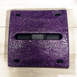 May include: A purple glitter-covered square-shaped box with a black rectangular opening in the center. The box has two black rectangular tabs on each side.