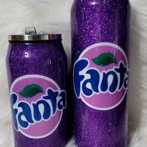 Puede incluir: Dos vasos aislados de purpurina morada con un logotipo "Fanta" rosa y azul. Los vasos tienen forma de lata.