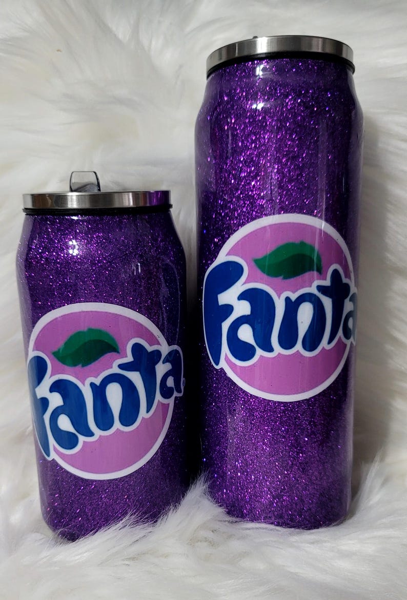 Puede incluir: Dos vasos aislados de purpurina morada con un logotipo "Fanta" rosa y azul. Los vasos tienen forma de lata.