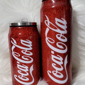 Puede incluir: Dos vasos t&eacute;rmicos rojos con purpurina con un logotipo blanco de Coca-Cola. Los vasos tienen forma de lata.