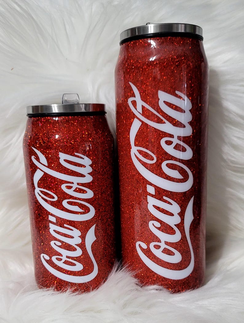 Puede incluir: Dos vasos t&eacute;rmicos rojos con purpurina con un logotipo blanco de Coca-Cola. Los vasos tienen forma de lata.