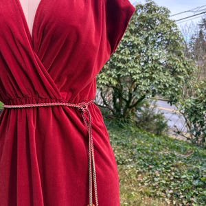 May include: A deep red, vintage-style dress with a V-neckline and short sleeves. The dress features a cinched waist and a decorative belt with two amber-colored beads. The dress is made of a soft, flowing fabric.