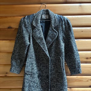 May include: A black and white tweed coat with a double-breasted design. The coat features a notched collar, two front pockets, and black leather shoulder accents. The coat is hanging on a metal hanger against a wooden background.