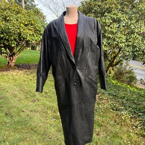 May include: A long, black leather coat with a notched collar and a single-button closure. The coat is displayed on a mannequin, revealing a red top underneath. The coat has long sleeves and a knee-length hemline. The background features green foliage.