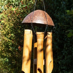 May include: A bamboo wind chime with a coconut shell top. The chime features several bamboo tubes of varying lengths, suspended by black cords. The tubes have angled cuts at the bottom. The background is a blurred view of green foliage.