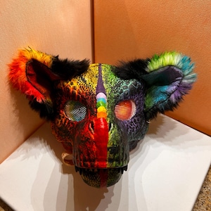 May include: A colorful, artistic mask with a bear-like design. The mask features a rainbow color scheme, with red, orange, yellow, green, blue, and purple hues. The ears are trimmed with black fur. The eyes are mesh, and the mouth has visible teeth.