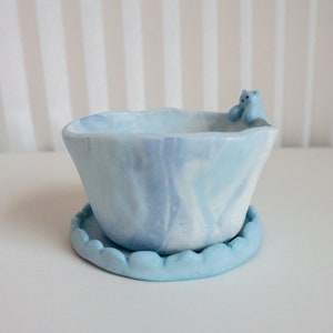 May include: A light blue ceramic planter with a marbled design and a small blue bear figurine on the rim. The planter has a matching saucer.