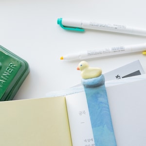 May include: A yellow duck bookmark with a blue and white watercolor design, placed in an open book. Two white highlighters with green and orange tips are visible. A green container is in the background.