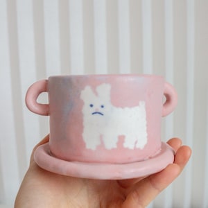 May include: A pink ceramic teacup and saucer set with a white dog illustration. The dog has a sad expression and is painted in a simple style.