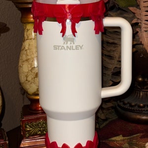 May include: White travel mug with a red bow design around the top and bottom. The mug has a handle and a straw lid. The mug has the word "STANLEY" printed on the side.