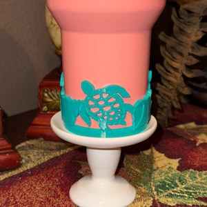May include: A pink ceramic tumbler with a teal sea turtle design around the rim, sitting on a white pedestal cake stand.