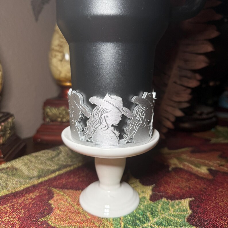 May include: A black tumbler with a silver decorative band featuring a silhouette of a woman wearing a cowboy hat. The tumbler sits on a white pedestal base. The design is detailed and the overall aesthetic is rustic.
