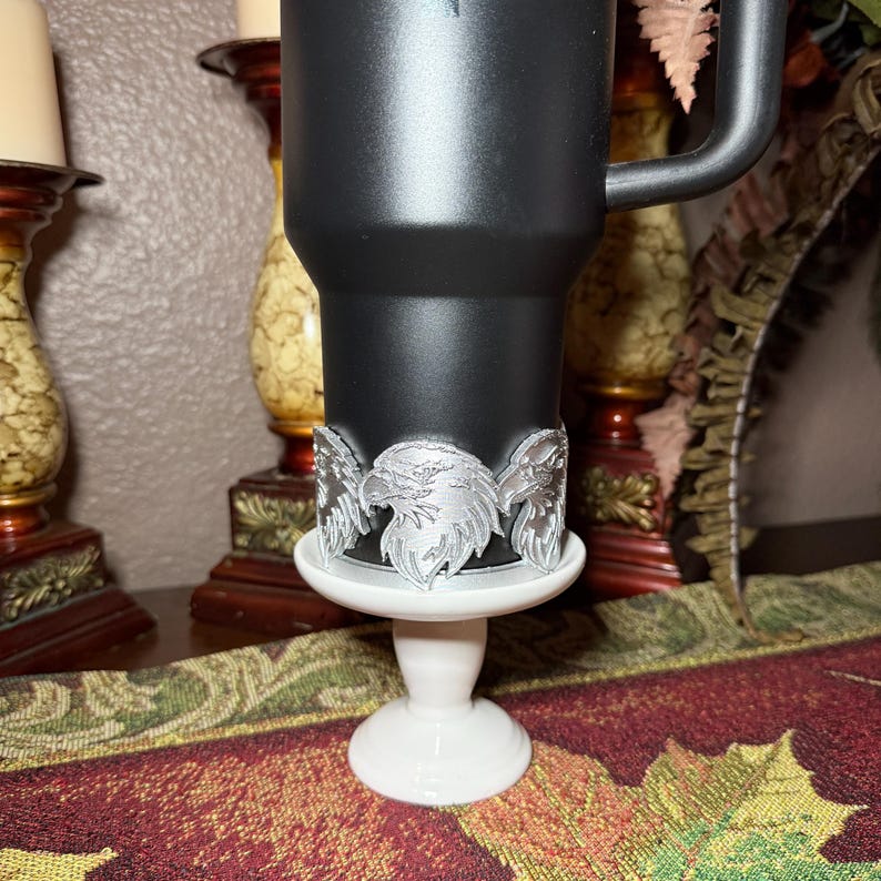 May include: A black tumbler with a silver eagle design, resting on a white pedestal. The eagle design wraps around the tumbler. The tumbler has a handle. The pedestal is white and has a round base.