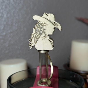 May include: A gold-colored bottle stopper featuring a detailed silhouette of a person wearing a cowboy hat. The stopper has a clear glass tube and a gold-colored ring. The design is intricate and stands out against the background.