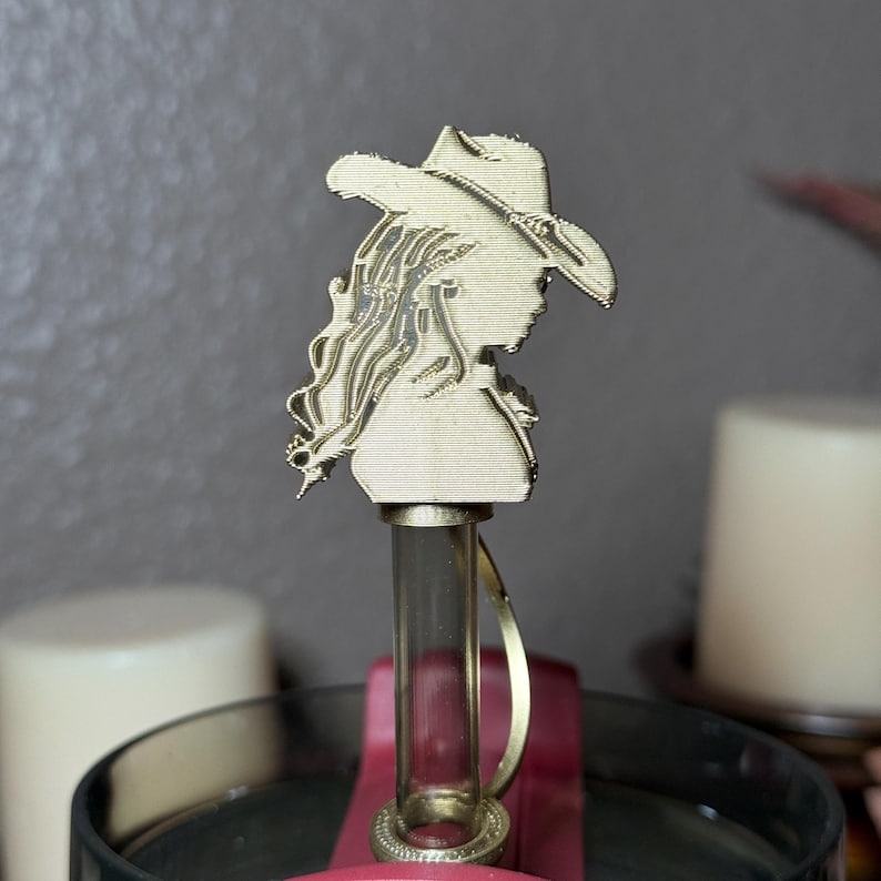 May include: A gold-colored bottle stopper featuring a detailed silhouette of a person wearing a cowboy hat. The stopper has a clear glass tube and a gold-colored ring. The design is intricate and stands out against the background.