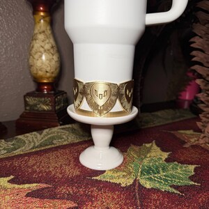 May include: A gold-colored metal cuff bracelet with a shield design, resting on a white pedestal. The bracelet is wrapped around a white mug. The background includes a decorative table runner and a candlestick.