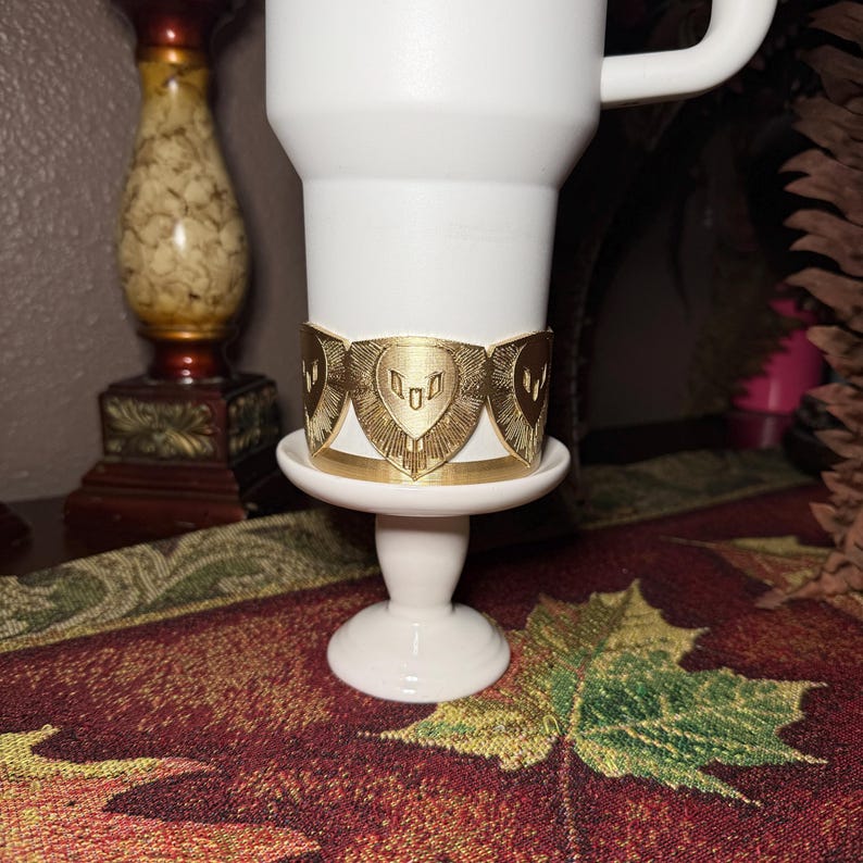 May include: A gold-colored metal cuff bracelet with a shield design, resting on a white pedestal. The bracelet is wrapped around a white mug. The background includes a decorative table runner and a candlestick.