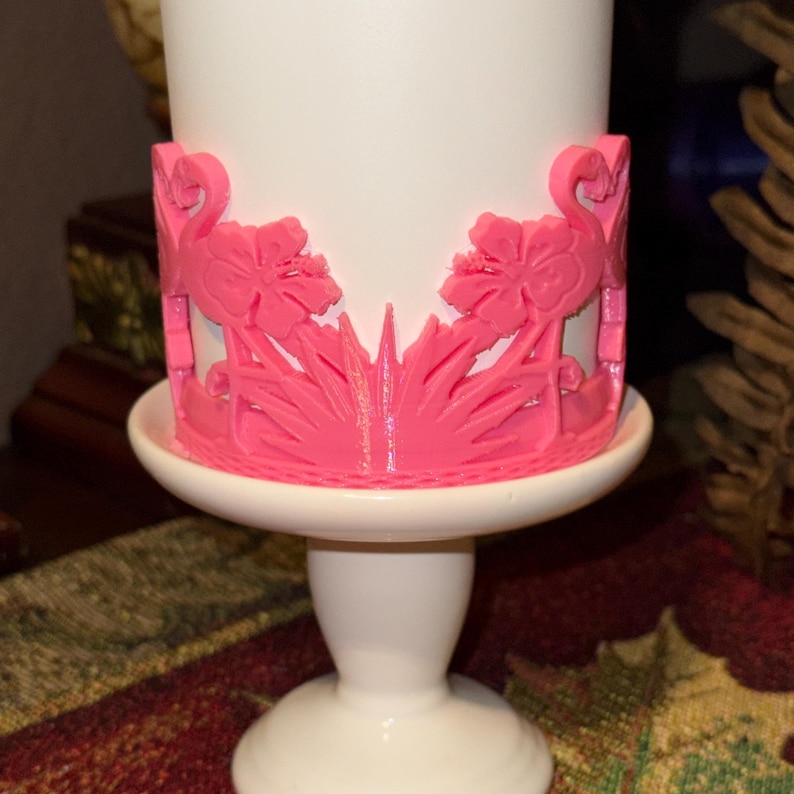May include: A white pedestal stand featuring a bright pink decorative wrap. The wrap displays a repeating pattern of stylized flamingos and hibiscus flowers, with a sunburst design at the center, encircling the base of the white cylindrical object.