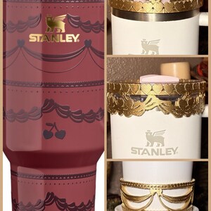 May include: A maroon Stanley tumbler with a decorative design, featuring cherries and a gold logo. The image also shows white Stanley tumblers with gold decorative bands, including heart and bow designs.