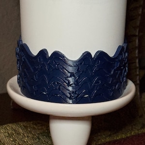 May include: A dark blue, flexible band featuring a repeating pattern of embossed stars and wavy lines, wrapped around a plain white cylindrical object. The band has a scalloped top edge and sits on a white, circular, pedestal-style base.