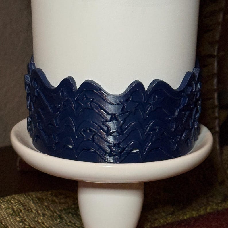 May include: A dark blue, flexible band featuring a repeating pattern of embossed stars and wavy lines, wrapped around a plain white cylindrical object. The band has a scalloped top edge and sits on a white, circular, pedestal-style base.
