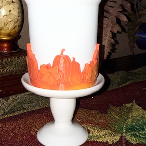 May include: A white ceramic candle holder with an orange pumpkin-themed design around the base. The holder sits on a white pedestal base, perfect for fall decor. The pumpkin design includes various pumpkin shapes and leaves.
