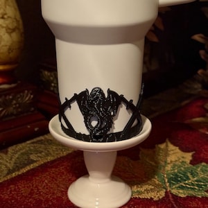 May include: A white ceramic mug with a black 3D printed dragon design around the middle. The mug is sitting on a white ceramic pedestal.