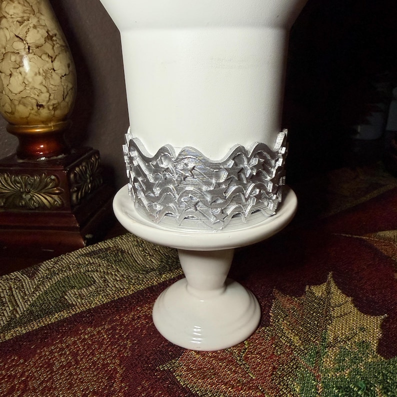 May include: A white ceramic pedestal vase featuring a decorative silver-colored band wrapped around its center. The band displays a repeating pattern of wavy lines and star shapes, creating a textured metallic contrast against the smooth white finish.