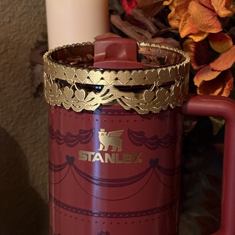May include: A red Stanley mug with a gold-colored decorative rim featuring heart and leaf designs. The mug has a handle and a lid. The Stanley logo is visible on the front of the mug. The mug has a decorative pattern.