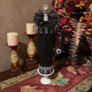 May include: Black tumbler with decorative embellishments. Features a jeweled bow, dangling rhinestones, and a crystal topper. The tumbler sits on a white pedestal base and has a black handle. The design includes black feathers and additional rhinestones.