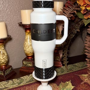 May include: White and black insulated tumbler with a handle. The tumbler has the word "COLDEST" printed on it. The lid and base have a textured design. The tumbler is on a white base.