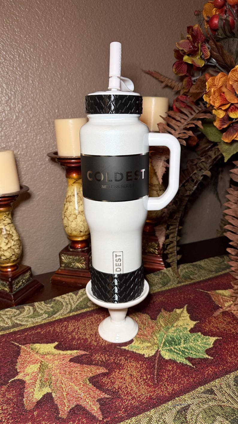 May include: White and black insulated tumbler with a handle. The tumbler has the word "COLDEST" printed on it. The lid and base have a textured design. The tumbler is on a white base.