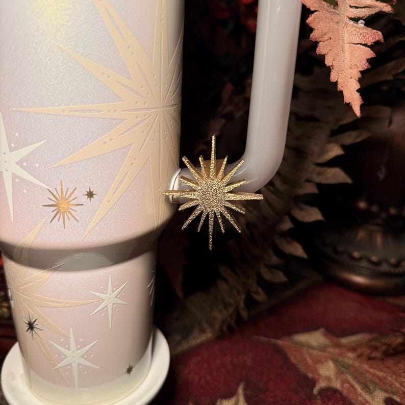 May include: A light pink and white travel mug with a starburst design and a gold-coloured star-shaped charm attached to the handle. The mug sits on a white saucer.
