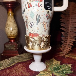 K&ouml;nnte beinhalten: Ein wei&szlig;er Reisebecher mit einem Western-Design mit Kakteen, Hufeisen und einem Longhorn. Der Becher steht auf einem wei&szlig;en Sockel mit einem goldfarbenen Zierrand. Der Becher hat einen wei&szlig;en Griff und ein Etikett mit der Aufschrift "Dishwasher Safe Traveler 40oz."