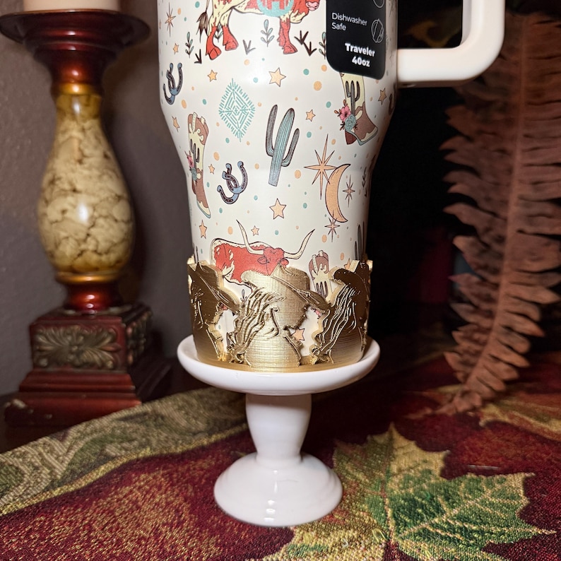 K&ouml;nnte beinhalten: Ein wei&szlig;er Reisebecher mit einem Western-Design mit Kakteen, Hufeisen und einem Longhorn. Der Becher steht auf einem wei&szlig;en Sockel mit einem goldfarbenen Zierrand. Der Becher hat einen wei&szlig;en Griff und ein Etikett mit der Aufschrift "Dishwasher Safe Traveler 40oz."