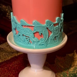 May include: A pink candle with a turquoise blue decorative trim featuring a dolphin and sea turtle design. The candle is on a white pedestal.