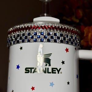 May include: White Stanley mug with a decorative rim featuring red, blue, and clear rhinestones. The mug is adorned with red, blue, and black star decals. The Stanley logo is prominently displayed on the front.