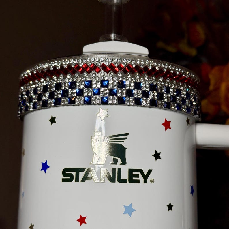 May include: White Stanley mug with a decorative rim featuring red, blue, and clear rhinestones. The mug is adorned with red, blue, and black star decals. The Stanley logo is prominently displayed on the front.