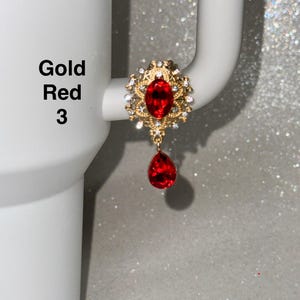May include: A gold-colored earring with a large, oval, red gemstone at the center, surrounded by smaller clear stones. A teardrop-shaped red gemstone hangs below. The text "Gold Red 3" is visible.