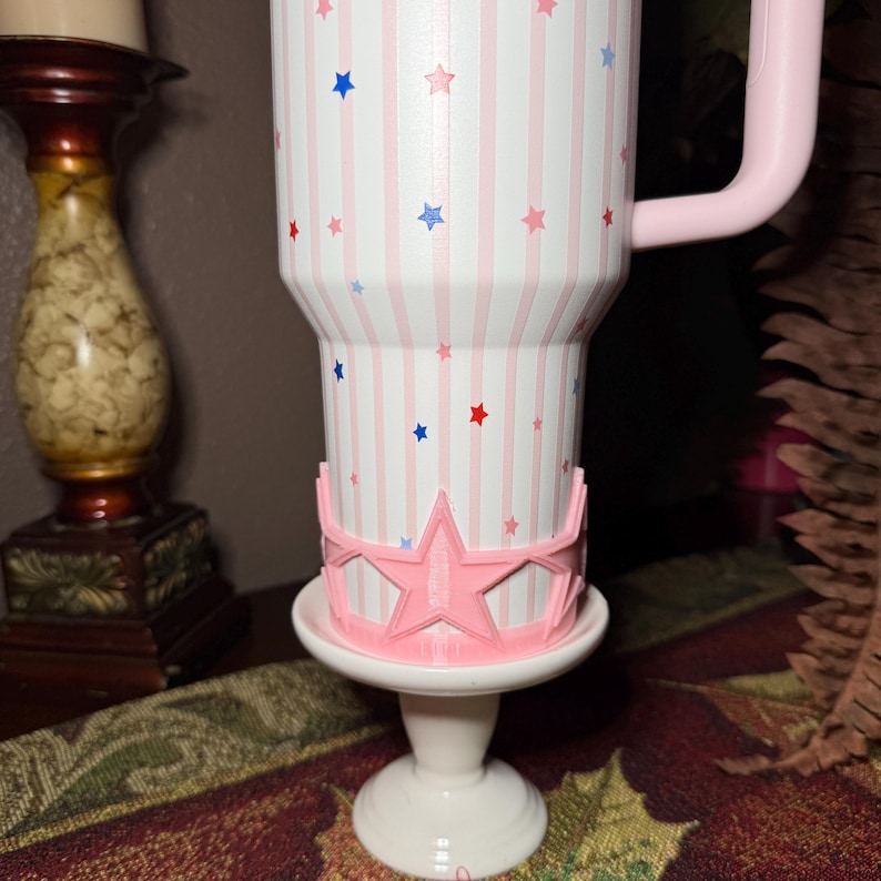 May include: A white tumbler with pink stripes and red, white, and blue stars. The tumbler has a pink handle and sits on a white pedestal with a pink star-shaped crown. The tumbler is a drinkware item.