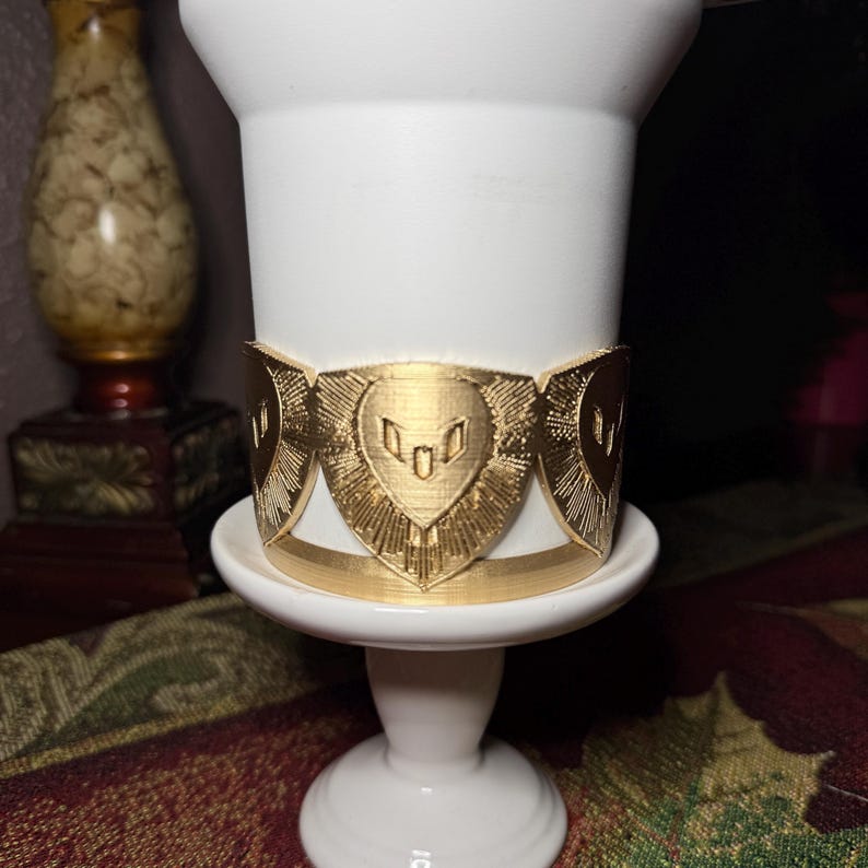 May include: A gold-colored decorative band with a heart-shaped design encircles a white, cylindrical object. The band features a repeating pattern of stylized hearts and geometric shapes. The object sits on a white pedestal.