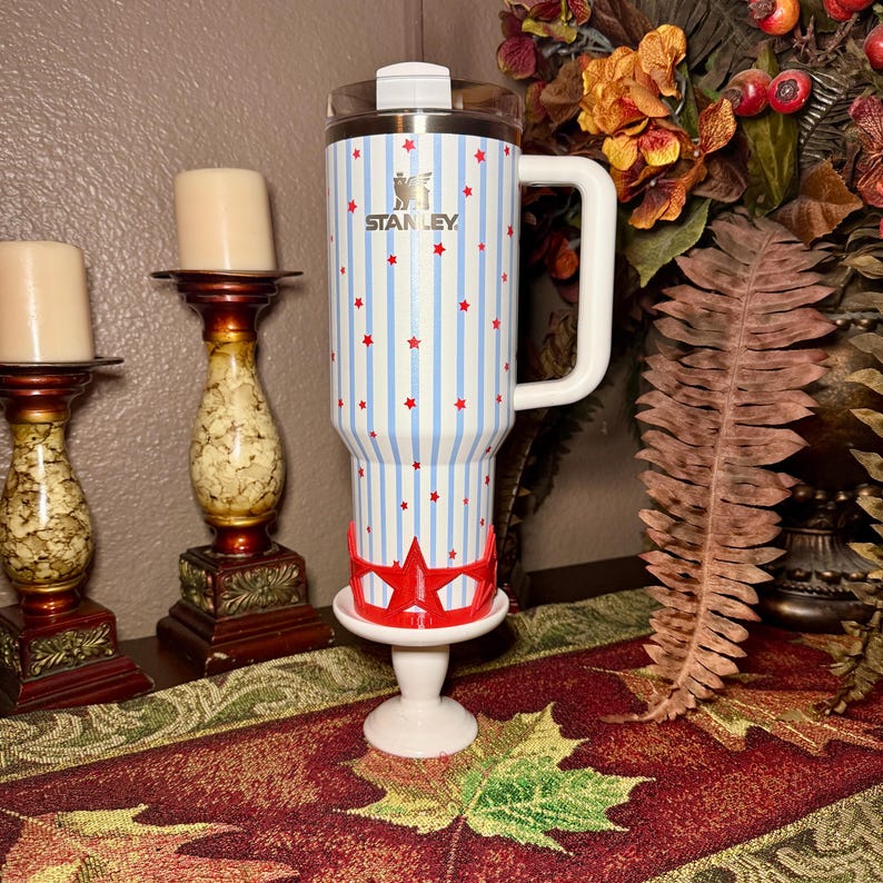 May include: A Stanley tumbler with a white handle, adorned with blue vertical stripes and red star accents. The tumbler is displayed on a white pedestal with a red star-shaped base. The Stanley logo is visible.